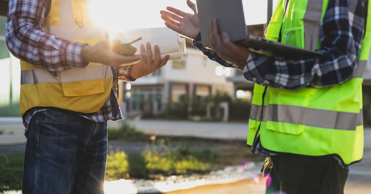 Two men in flannel shirts and high-visibility vests are talking, with one holding rolls of paper and the other a tablet.