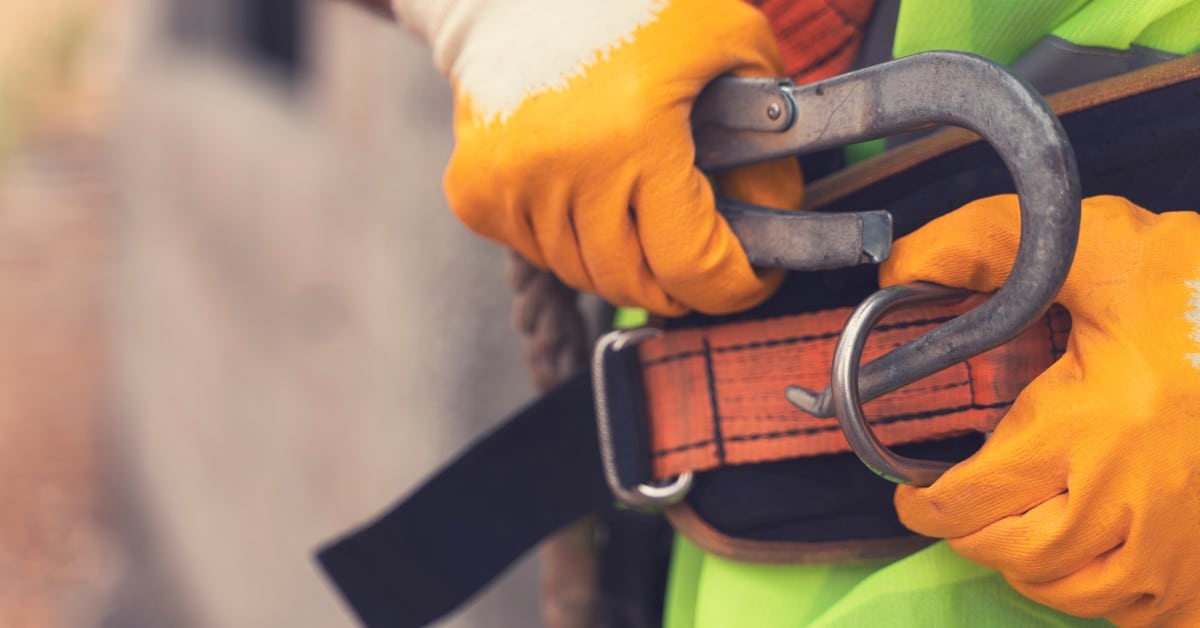 A close-up of a worker with orange gloves and a high-visibility vest hooking on a harness to a safety belt.