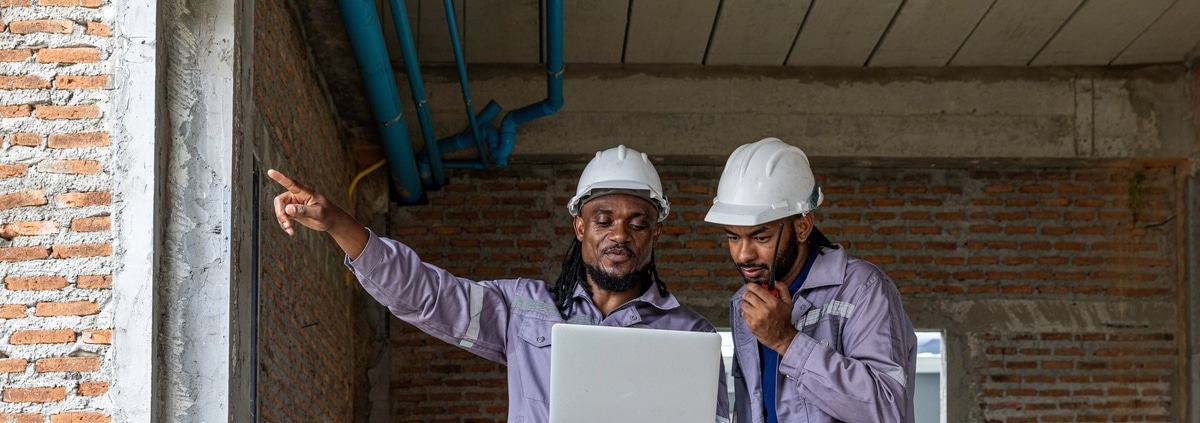 Two workers in hard hats and purple work shirts look at a laptop on a construction site while one talks on a walkie-talkie.