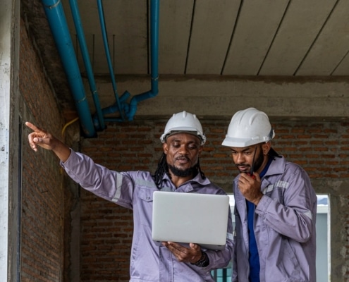 Two workers in hard hats and purple work shirts look at a laptop on a construction site while one talks on a walkie-talkie.