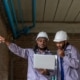 Two workers in hard hats and purple work shirts look at a laptop on a construction site while one talks on a walkie-talkie.