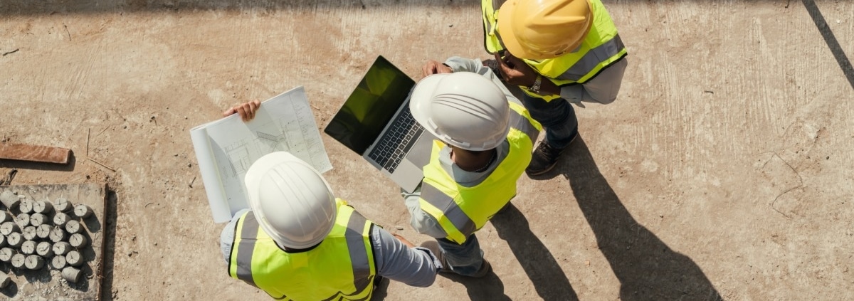 An overhead view of three construction workers in high-visibility vests and hard hats talking on a construction site.