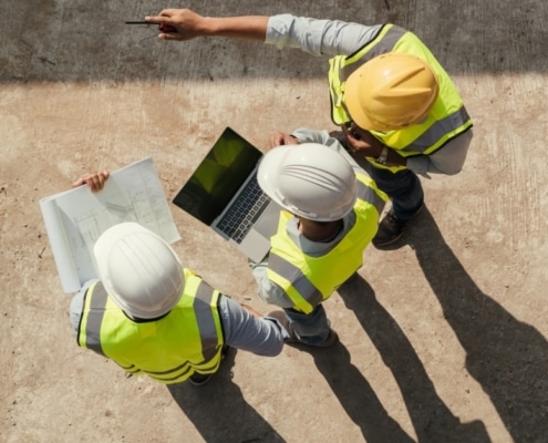 An overhead view of three construction workers in high-visibility vests and hard hats talking on a construction site.