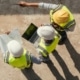 An overhead view of three construction workers in high-visibility vests and hard hats talking on a construction site.