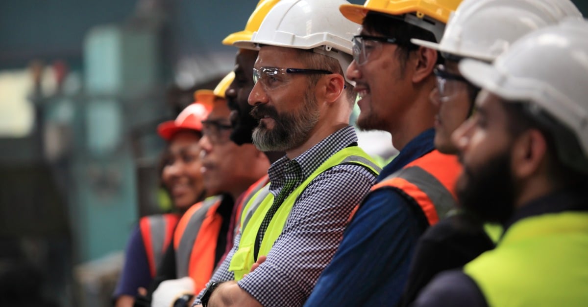 A line of eight construction workers in high-visibility vests, hard hats, and safety goggles standing together.