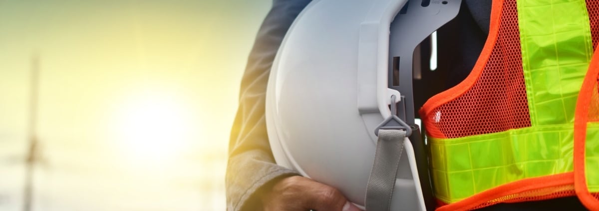 A close-up of a man with a high-visibility vest holding a white hard hat under his arm close to his waist.