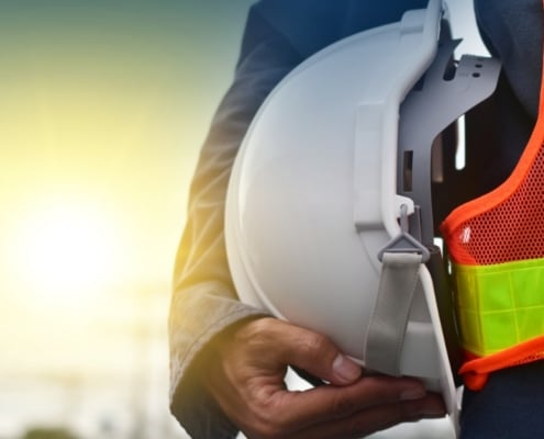 A close-up of a man with a high-visibility vest holding a white hard hat under his arm close to his waist.