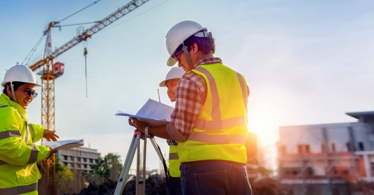 Three workers in high-visibility apparel and hard hats talk with folders and documents on a construction site.
