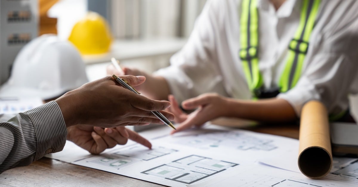 A close-up of a woman's hands with a pen pointing at blueprints on a desk, with another person across from them.