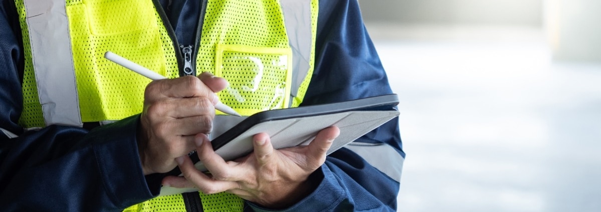 A close-up of a man in a work jumpsuit with a high visibility vest, making notes with a white pen on an electronic tablet.