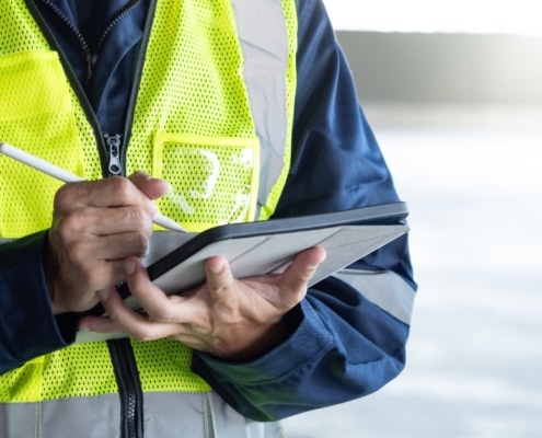 A close-up of a man in a work jumpsuit with a high visibility vest, making notes with a white pen on an electronic tablet.