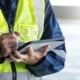 A close-up of a man in a work jumpsuit with a high visibility vest, making notes with a white pen on an electronic tablet.