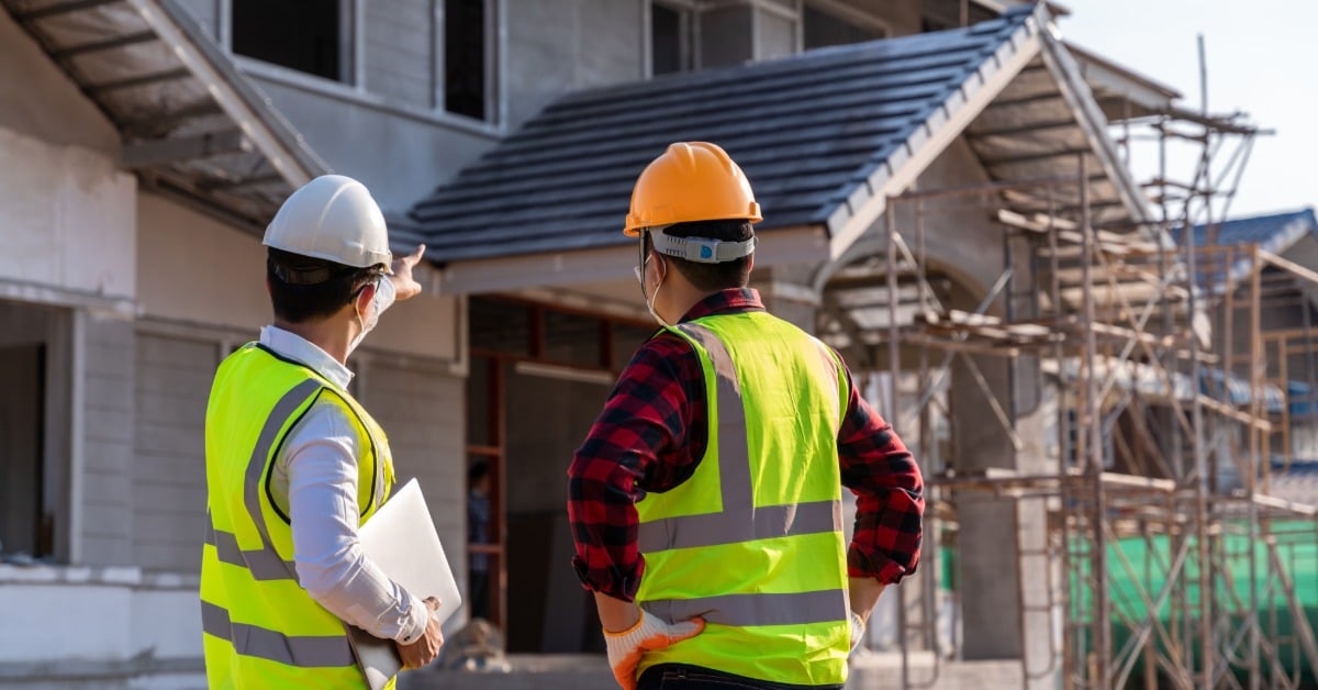 Two workers in high-visibility vests and a hard hat, with one holding a clipboard, talking while on a construction site.