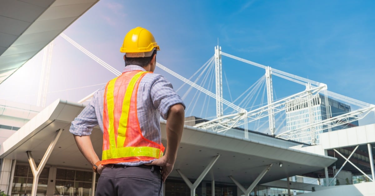A view from behind of a man in a high-visibility vest and a hard hat looking at a building's construction site.