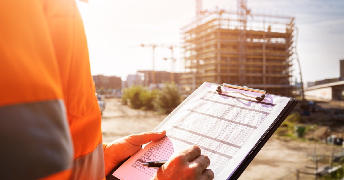 A close-up of a man in an orange, high-visibility jacket checking boxes on a sheet on a clipboard at a construction site.