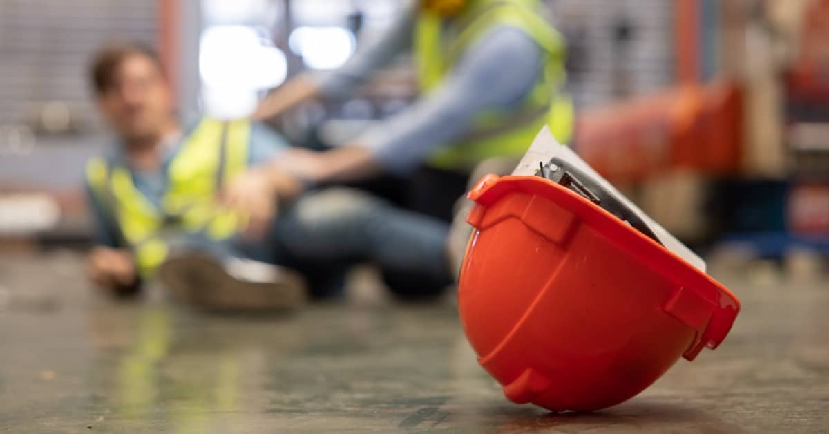 A close-up of an overturned hard hat on the floor, with a blurred injured worker and another helping him in the background.