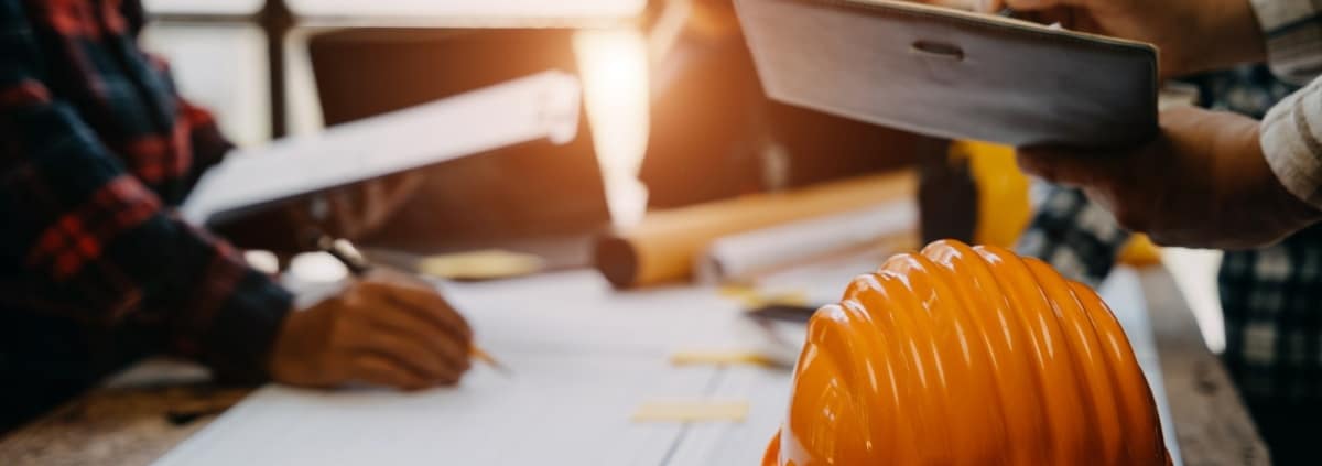 A close-up of an orange hard hat on a wooden table with people having a meeting with documents in the background.