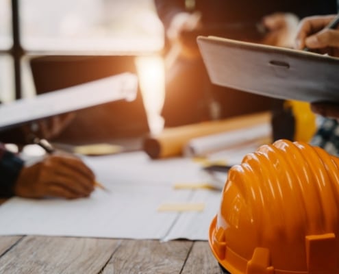 A close-up of an orange hard hat on a wooden table with people having a meeting with documents in the background.