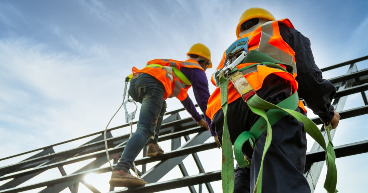 A rear view of two workers with high-visibility vests climbing a roof structure while wearing a fall arrestor device.