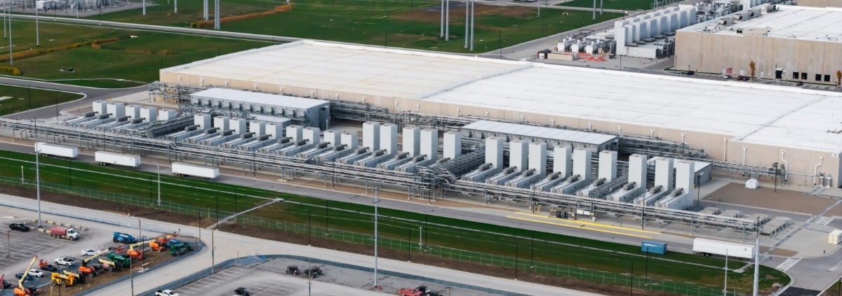 An aerial view of a large data center campus during the day with numerous cooling units, grass fields, and parking lots.