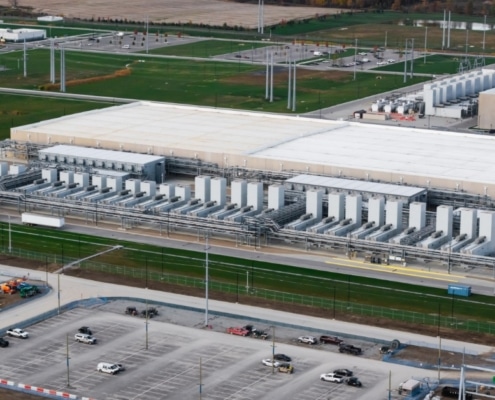 An aerial view of a large data center campus during the day with numerous cooling units, grass fields, and parking lots.