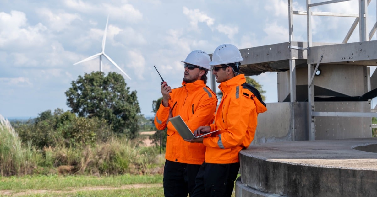 Two men in orange jackets outside near a large infrastructure. One talks on a walkie-talkie while the other uses a laptop.
