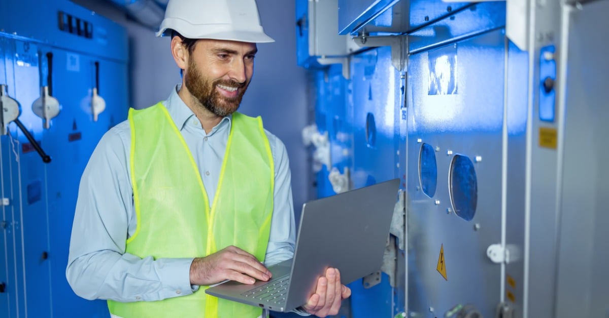 A man with a white hard hat, a high-visibility vest, and a laptop smiles as he inspects something in a data center.