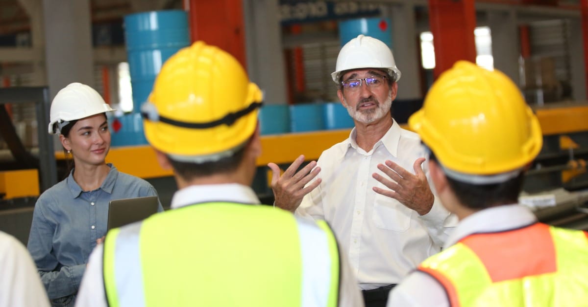 A line of people in hard hats and high-visibility vests listening to a man and woman in business attire in hard hats talk.