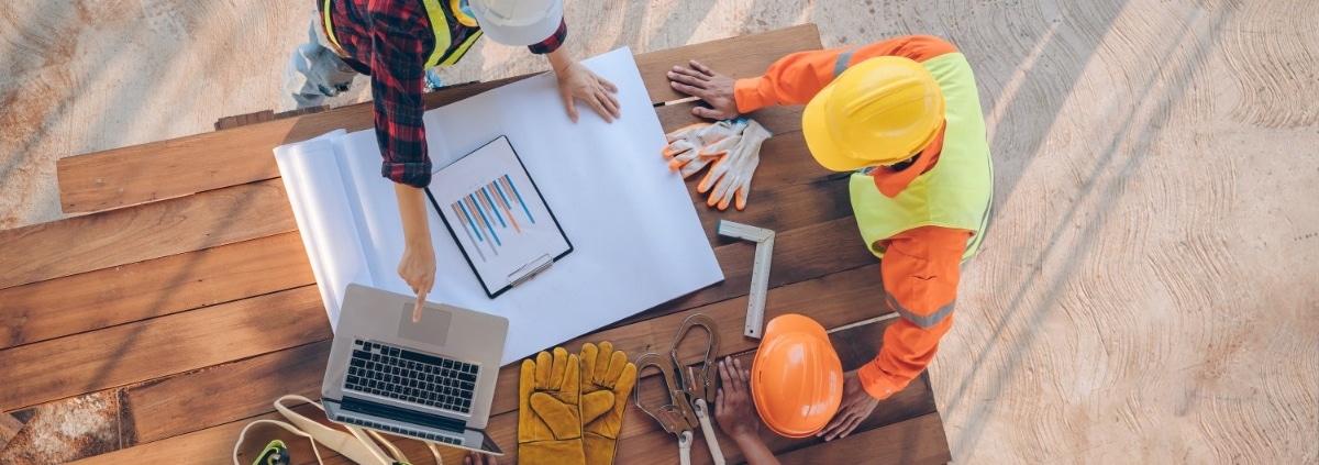 An overhead view of three construction workers in hard hats and vests leaning on a wooden table and discussing plans.