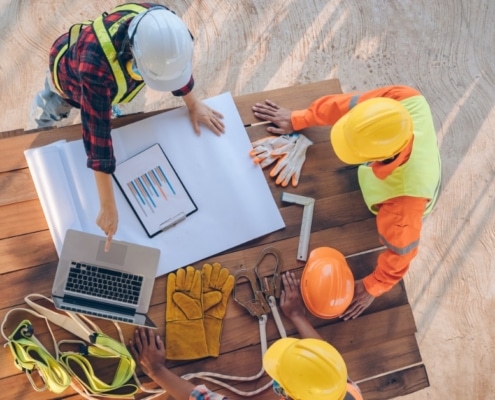 An overhead view of three construction workers in hard hats and vests leaning on a wooden table and discussing plans.