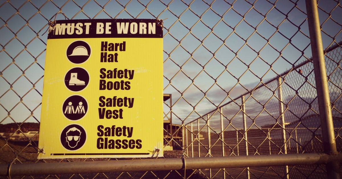 A yellow and black safety sign on a chain-link fence that details personal protective equipment requirements.