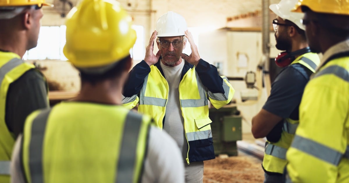 A group of people in hard hats and high-visibility vests huddle around a man in a hat and vest talking.
