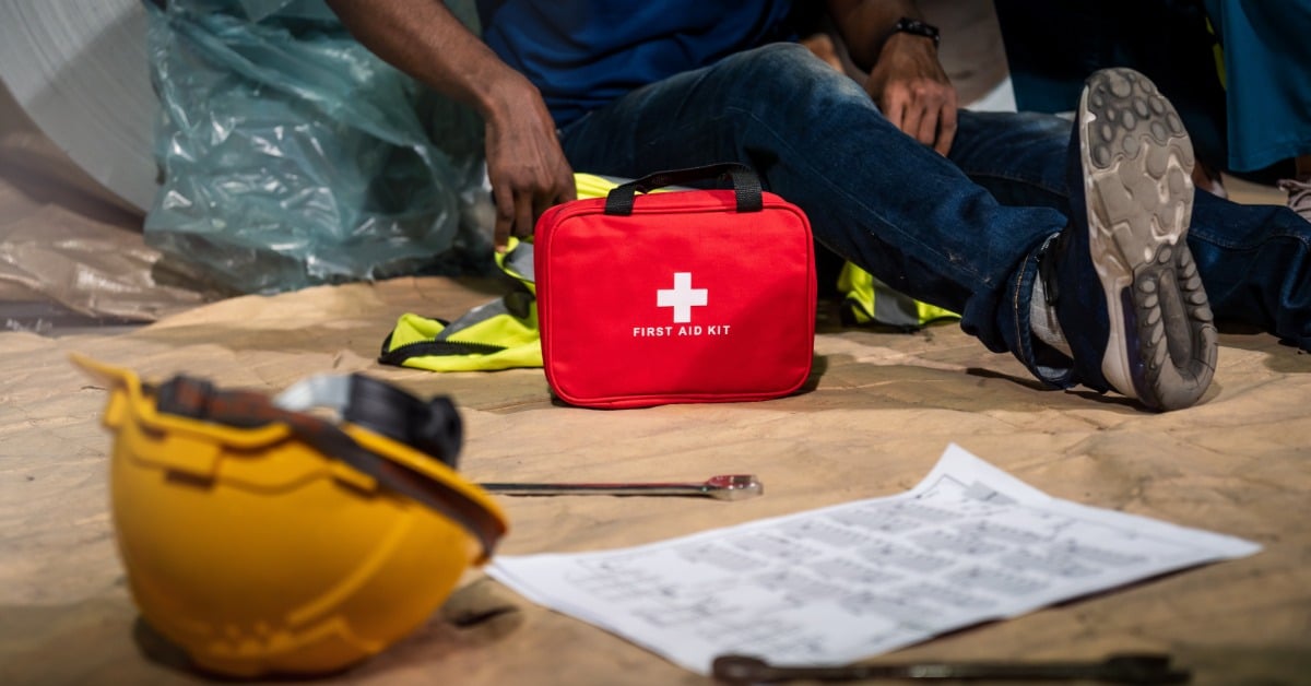 A close-up of a man's lower body as he sits on the ground next to a first aid kit and a yellow hard hat.