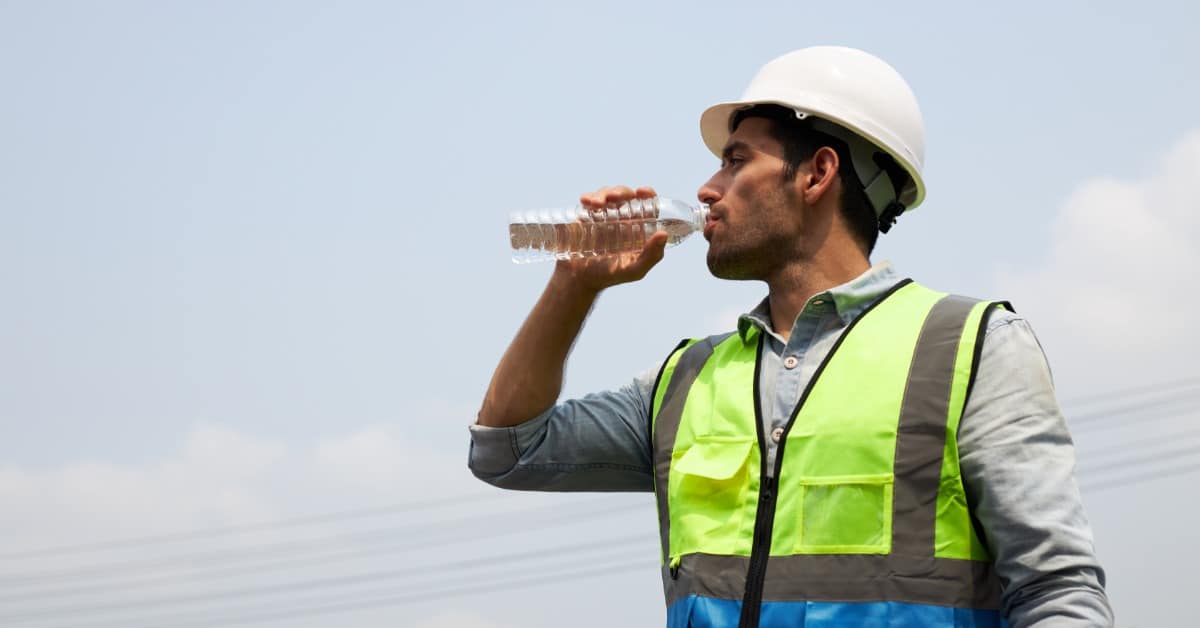 A man in a dress shirt and a high-visibility neon vest drinks water from a clear plastic bottle outside.