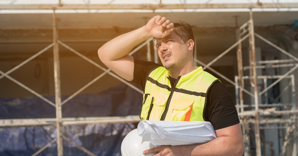 A man in a black t-shirt and high-visibility safety vest wipes his sweating brow while holding a white hard hat.