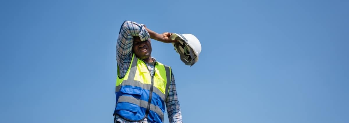 A man in a flannel shirt, jeans, and a high-visibility vest wipes his brow while holding a white hard hat.