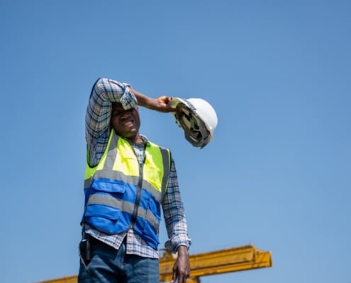 A man in a flannel shirt, jeans, and a high-visibility vest wipes his brow while holding a white hard hat.