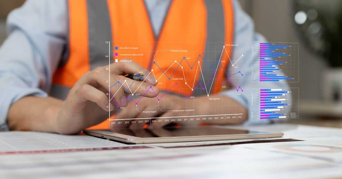 The torso of a man with an orange high-visibility vest at a desk with a holographic chart of data in front of him.