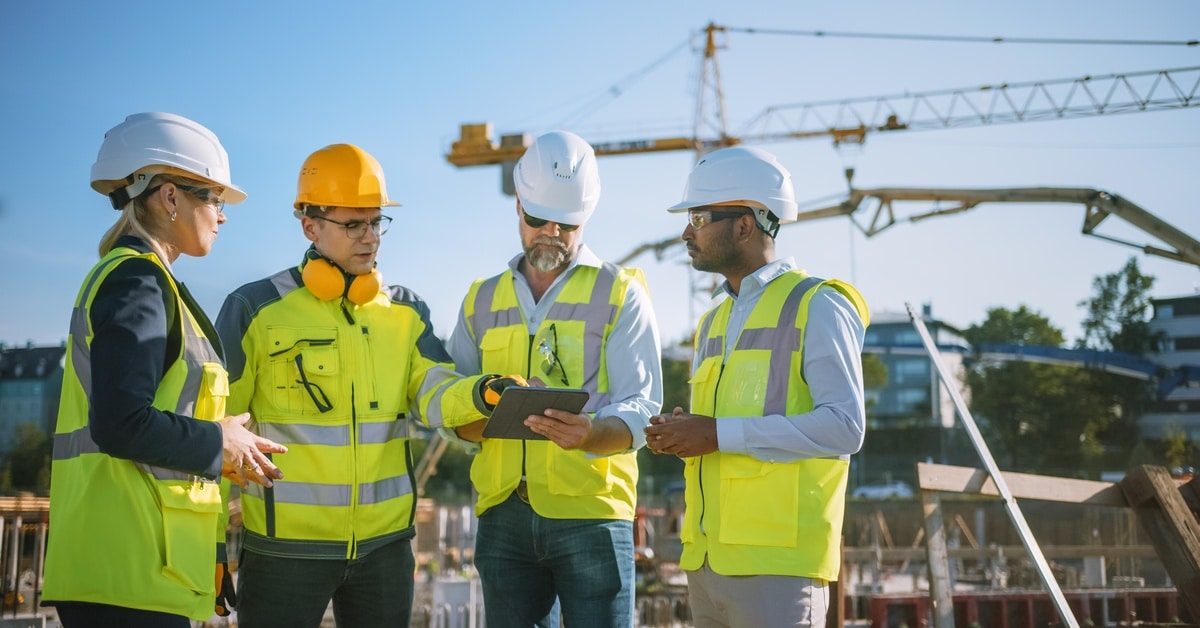 A group of four men and women in high-visibility safety vests with hard hats looking at an e-tablet on a construction site.
