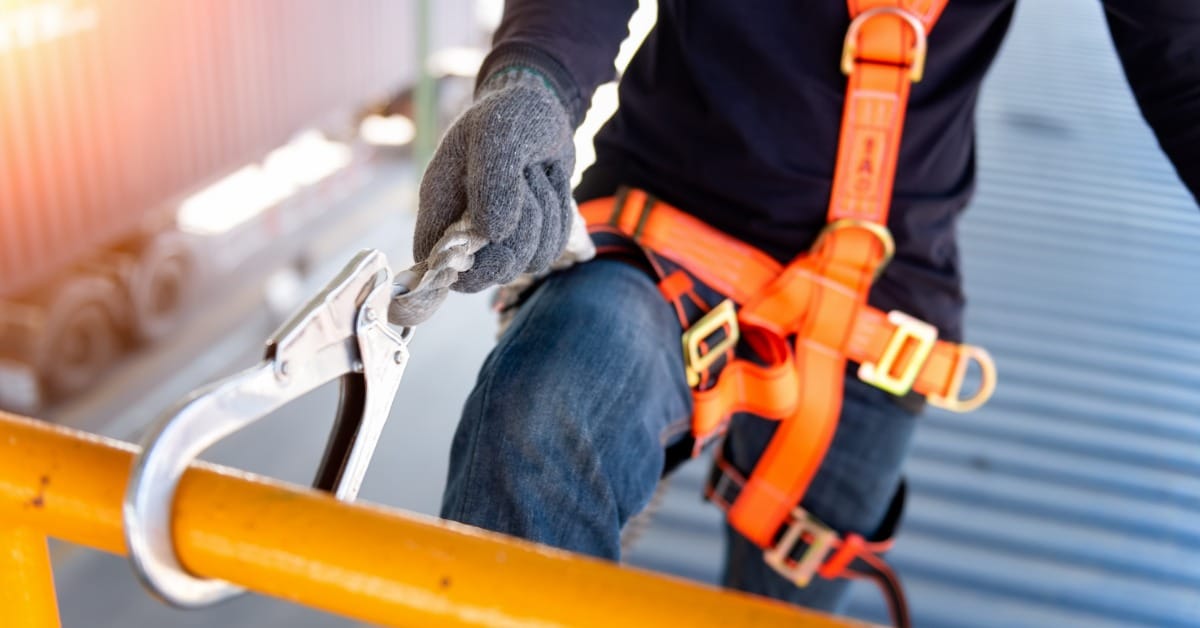 A close-up of a man in jeans, a black top, and gloves attaching the hook and line to his safety harness to a yellow railing.