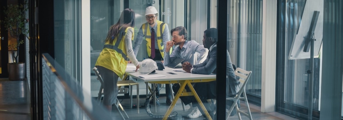A group of men and women, some in business attire, others in construction attire, talk in a glass conference room.