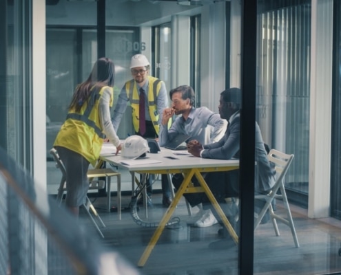A group of men and women, some in business attire, others in construction attire, talk in a glass conference room.