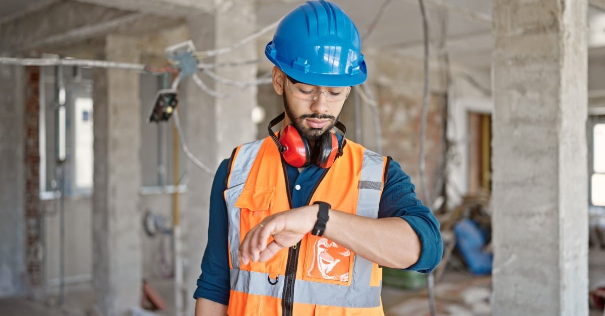 A man in a high-visibility vest, a blue hard hat, ear protection, and safety goggles checks his watch on a work site.