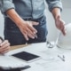 Three men at a desk table, gesturing demonstratively. On the table are construction plans, a hard hat, and clipboards.