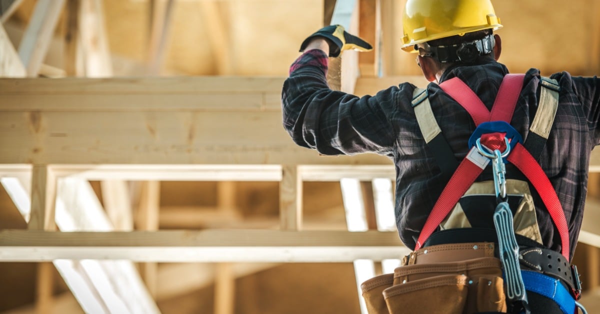 A view from behind of a construction worker with a hard hat, safety harness, and utility belt working on a wooden roof.