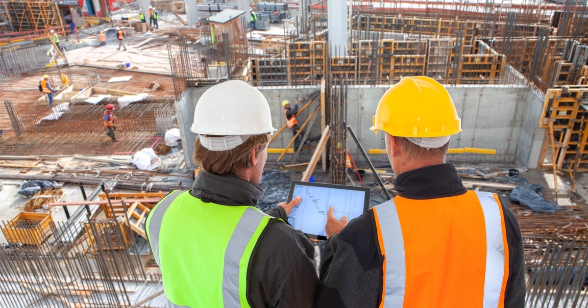 A rear view of two men in high-visbility vests and hard hats looking at an electronic tablet at a major construction site.