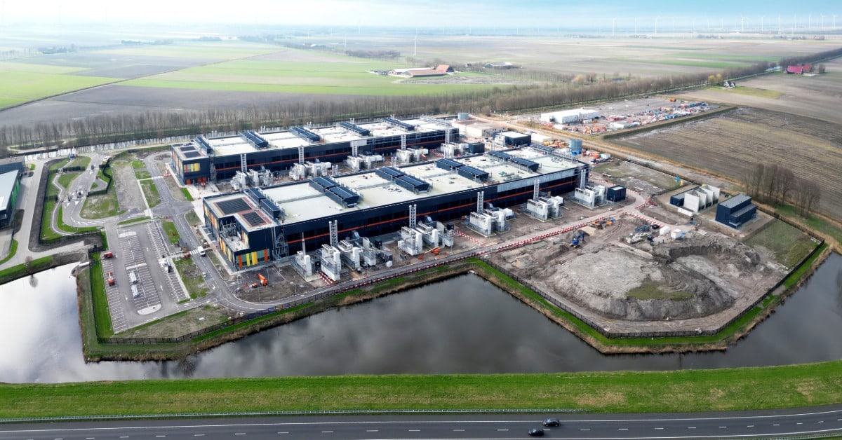 An aerial view of a large data center under construction and surrounded by water, with a road nearby on a sunny day.