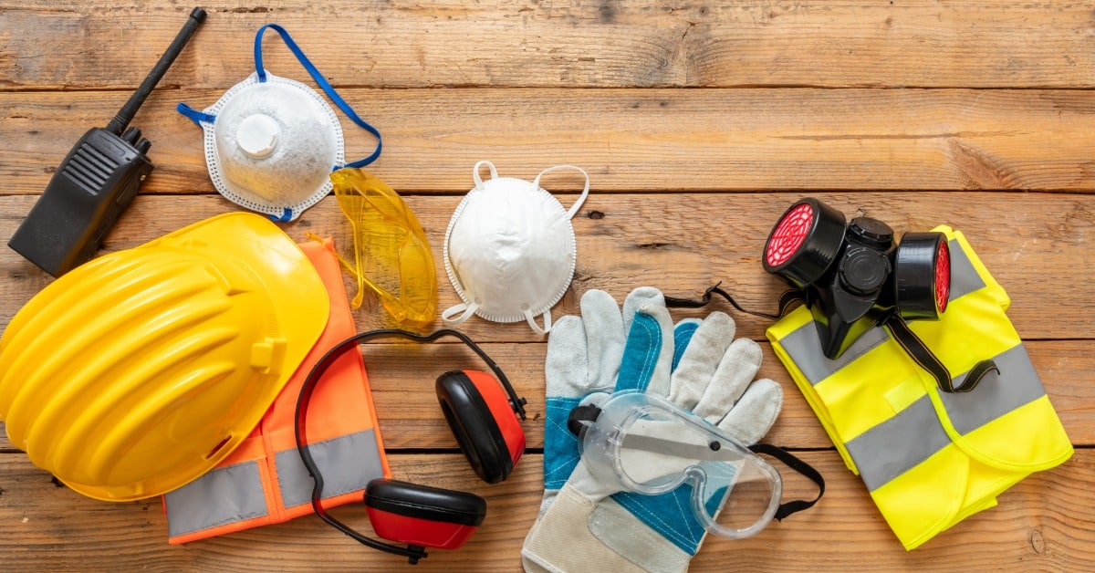 An overhead view of various construction safety equipment on a wooden table, including a hard hat, gloves, and masks.