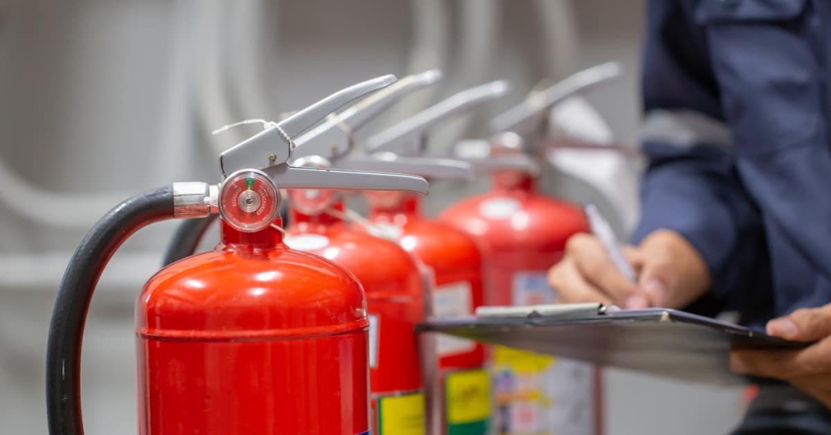 A close-up of a man writing on a blue clipboard in front of a row of four red fire extinguisher tanks.