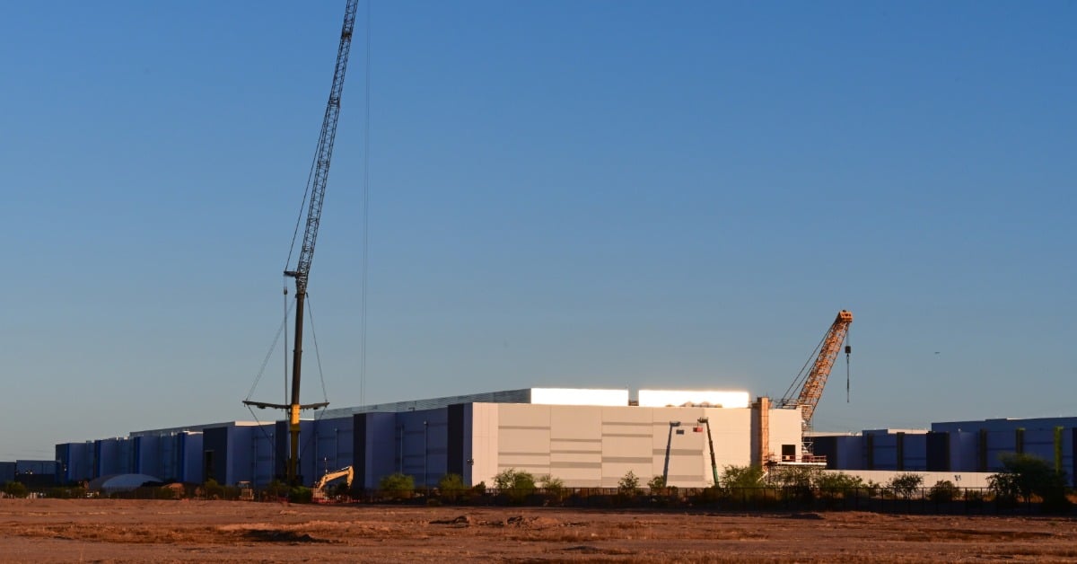 A wide landscape view of a large data center campus under construction with multiple large cranes and the sun setting.